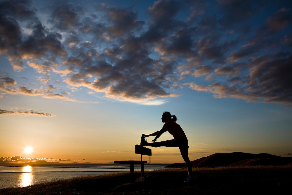 A woman stretches against a bench on a hill overlooking a bay at sunset.